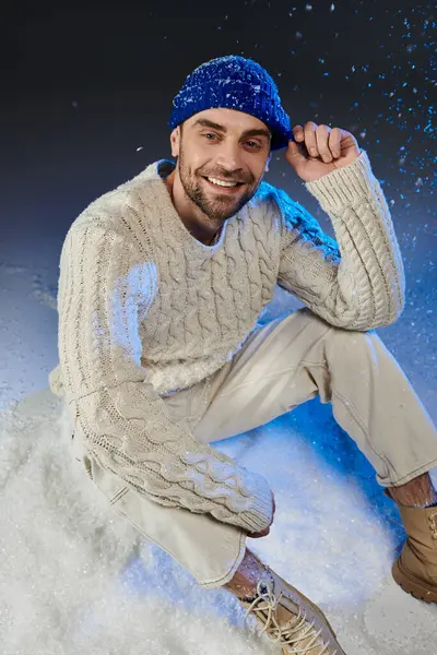 A cheerful young man sits in the snow, wearing a warm sweater and a blue hat, smiling happily. — Stock Photo