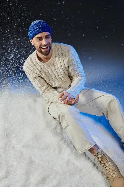 A cheerful young man sits on snow, enjoying the winter vibes while smiling in soft clothing. — Stock Photo