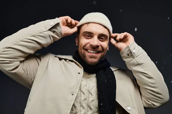 A cheerful young man smiles broadly while adjusting his warm hat in a winter setting with snow. — Stock Photo