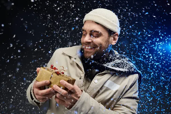 Um jovem bonito sorri amplamente, segurando um presente lindamente embrulhado em meio a neve girando. — Fotografia de Stock