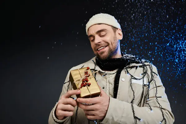 A cheerful young man embraces a golden gift while festive lights twinkle around him. — Stock Photo