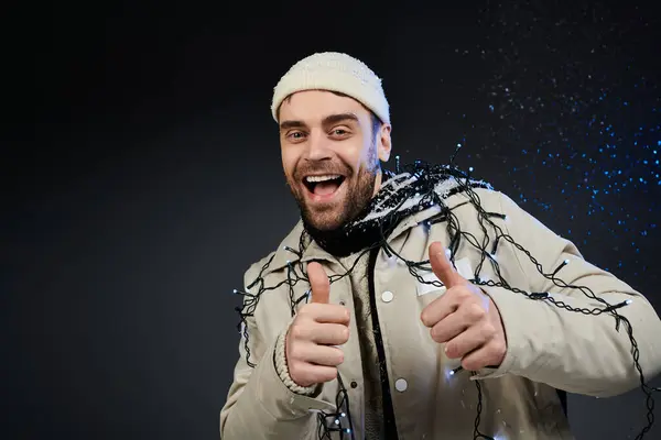 A cheerful young man gives two thumbs up while adorned in holiday lights and a cozy hat. — Stock Photo