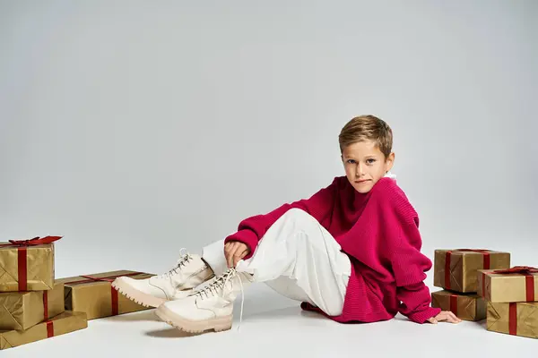 The boy sits comfortably in cozy winter attire, surrounded by beautifully wrapped gifts. — Stockfoto