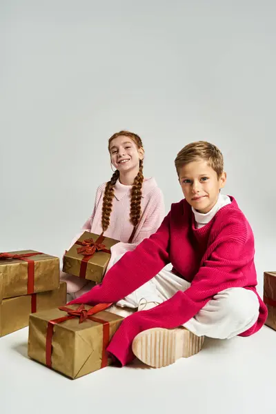 Two kids warmly dressed in festive clothing sit happily among beautifully wrapped presents. — Stock Photo