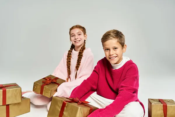 Two happy kids in warm clothing smile while sitting with colorful present boxes. — Stock Photo