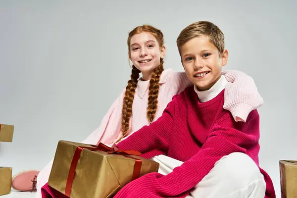 Two happy children in winter clothing sit together, surrounded by cheerful gift boxes during winter. — Stock Photo