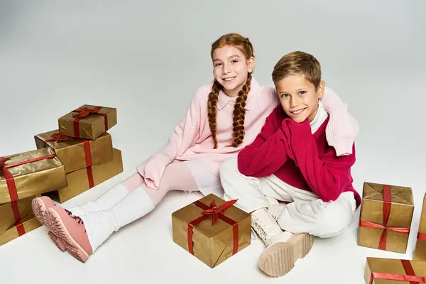 Two happy kids in warm attire enjoy the holiday spirit amid golden gifts with red ribbons. — Stock Photo