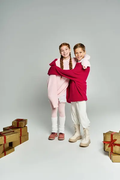 Two kids in cozy winter clothing embrace warmly, surrounded by beautifully wrapped presents. — Foto stock