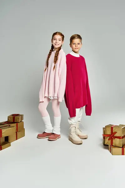 Two kids in playful winter outfits pose together amid wrapped presents, exuding holiday joy. — Stock Photo