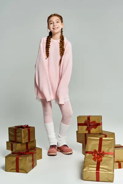 A joyful girl in cozy pink attire stands among golden wrapped gifts, radiating warmth and happiness. — Stock Photo