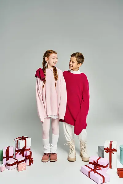 Two kids dressed in warm winter clothing are happily standing near colorful wrapped presents. — Stock Photo