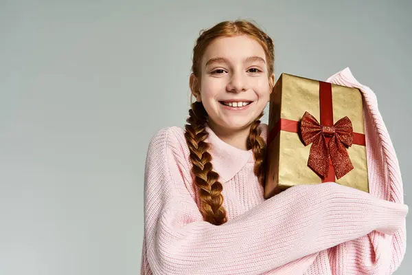 A cheerful girl dressed in cozy winter clothing beams while holding a golden gift with a bow. — Stock Photo