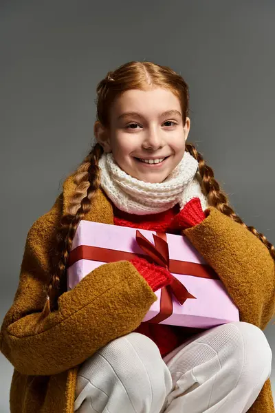 A cheerful young girl smiles warmly while embracing a colorful present in cozy winter clothing. — стоковое фото