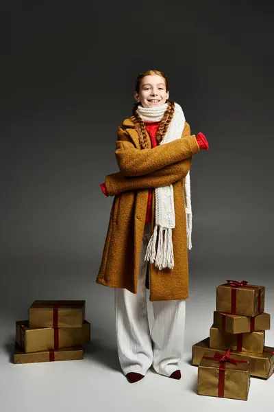 A cheerful youngster dressed in warm winter clothing stands by neatly stacked presents. — Stock Photo