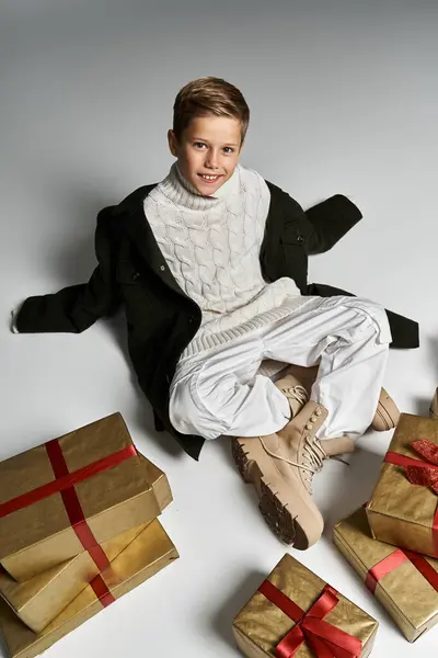 A young boy dressed warmly sits on the ground among beautifully wrapped gifts, radiating joy. — Stock Photo