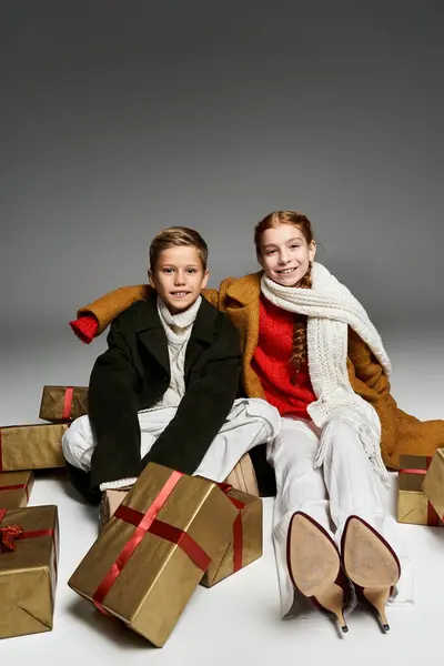 Two kids sitting in cozy winter clothes surrounded by gift boxes during the festive period. — Stock Photo