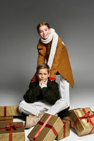 Two cheerful kids dressed warmly pose among beautifully wrapped gifts creating a joyful atmosphere. — Stock Photo