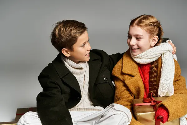 Two children in winter attire warmly interact while holding gifts surrounded by a cozy atmosphere. — Stock Photo