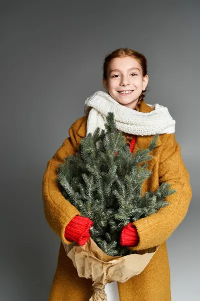 A girl in winter clothes joyfully shows a small Christmas tree, spreading cheer. — Stock Photo