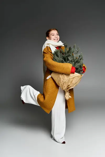 A girl dressed warmly is playfully holding a small Christmas tree, radiating joy. — Stock Photo