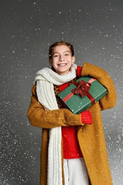 A delighted young girl dressed in warm winter clothes holds a gift while snowflakes fall around her. — Stock Photo