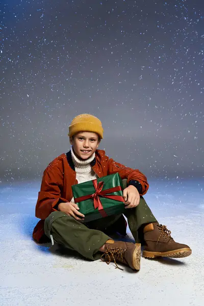 Ein kleiner Junge mit warmem Hut und Jacke genießt eine verschneite Umgebung, während er ein festliches Geschenk in der Hand hält. — Stockfoto