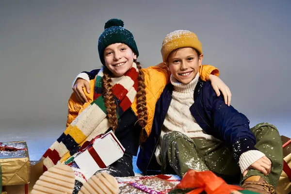 Two cheerful kids dressed warmly share smiles while surrounded by colorful presents and decorations. — Stock Photo