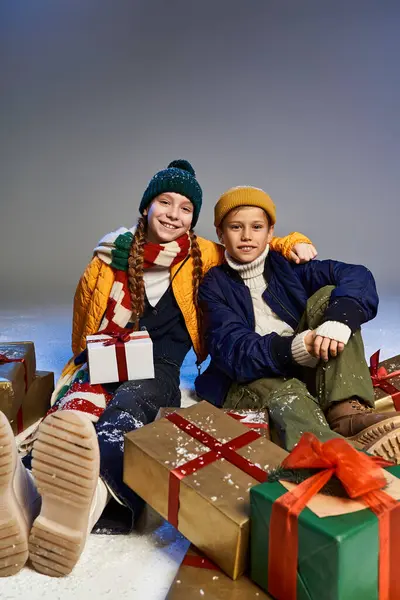 Two children dressed in warm winter attire sit on the floor next to wrapped Christmas presents. — Stock Photo