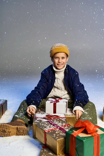A happy boy in winter gear sits among wrapped gifts in the falling snow. — Stock Photo