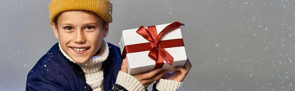 A cheerful young boy wearing warm layers smiles while presenting a beautifully wrapped gift. — Stock Photo