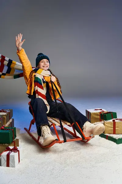 A happy girl dressed warmly enjoys sledding while waving amidst colorful presents. — Stock Photo