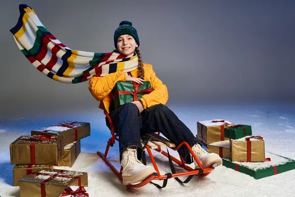 Un enfant joyeux enveloppé dans une écharpe colorée s'assoit sur un traîneau, entouré de cadeaux en hiver. — Photo de stock