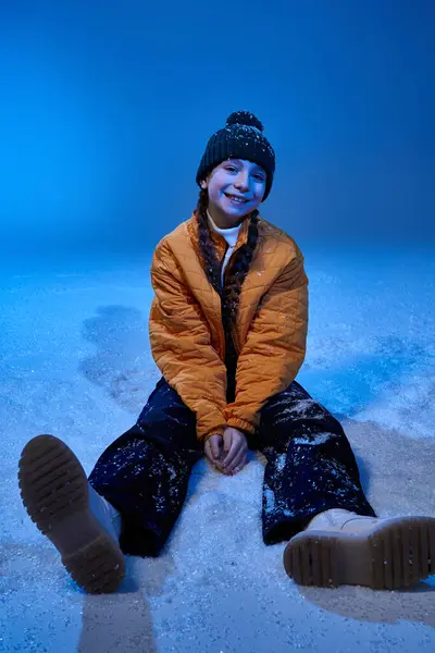A happy girl in cozy winter clothing sits on the snow, smiling brightly in a winter setting. — Stock Photo