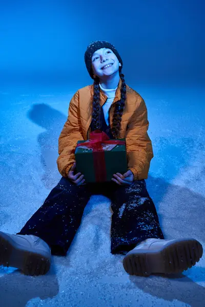 A girl with braids smiles warmly while seated in the snow, holding a beautifully wrapped gift. — Stock Photo