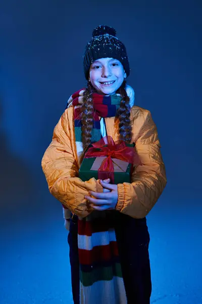 A young girl smiles happily, dressed warmly for winter, holding a beautifully wrapped present. — Stock Photo