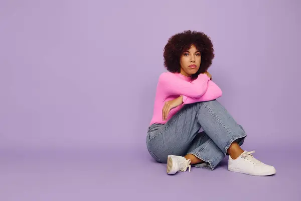 A young Black woman in pink sits thoughtfully, sitting against a vibrant purple background. — Stock Photo