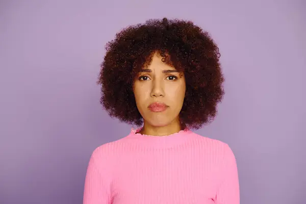 A young woman with curly hair showcases her sad emotion while wearing casual pink attire on a purple background. — Stock Photo