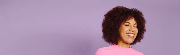 Eine fröhliche junge Frau mit lockigem Haar strahlt Glück aus, während sie lässig rosa Kleidung vor violettem Hintergrund trägt. — Stockfoto