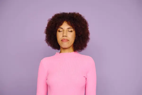 A young woman with curly hair, dressed in pink, showcases her emotions with closed eyes on a vibrant purple backdrop. — Stock Photo