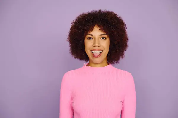 A young woman in pink clothing playfully sticks her tongue out, radiating joy against a purple background. — Stock Photo