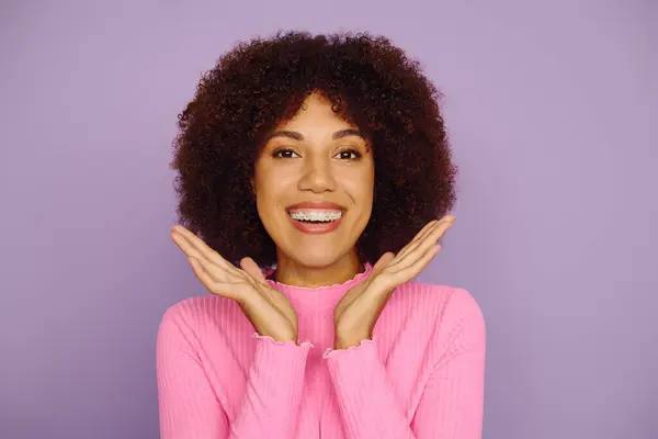 A young African American woman beams with joy, showcasing her curly hair while dressed in casual pink clothing. — Stock Photo