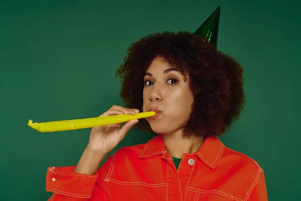Una giovane donna vivace con i capelli ricci esprime felicità, soffiando un corno di festa su uno sfondo verde vibrante. — Foto stock