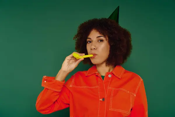 Una giovane donna allegra con i capelli ricci gode di un momento giocoso con un corno da festa, vestita con disinvoltura contro il verde. — Foto stock