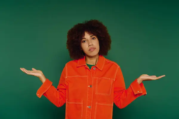 A cheerful young woman with curly hair showcases a shrug gesture while dressed casually on a green backdrop. — Stock Photo