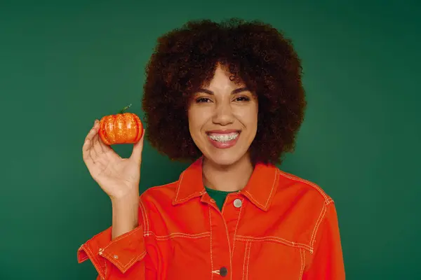 A cheerful young woman with curly hair expresses joy while holding a bright pumpkin in front of a green wall. — Stock Photo