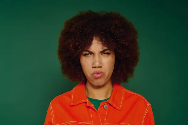 Una mujer afroamericana bastante joven con el pelo rizado muestra su emoción insatisfecha en ropa casual sobre un fondo verde. - foto de stock