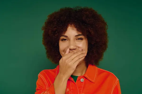A young woman with curly hair displays a joyful expression, her hand covering a smile on a green background. — Stock Photo