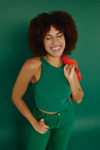 A young woman with curly hair radiates happiness in chic green casual wear, standing against a lively backdrop. — Stock Photo