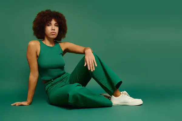 Una bella giovane donna afroamericana con i capelli ricci mette in mostra il suo stile mentre si siede su uno sfondo verde. — Foto stock
