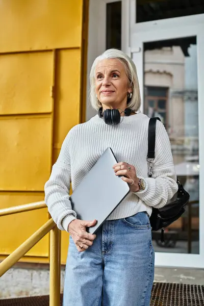 A confident gray haired woman smiles warmly, holding a laptop and wearing headphones. — Stock Photo